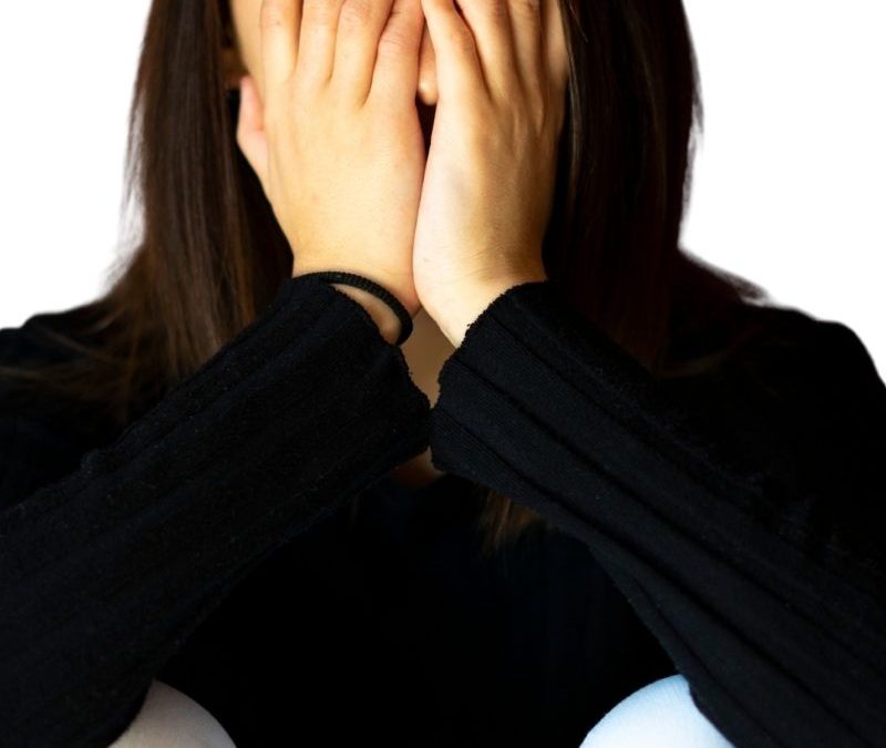 Woman with her hands covering her face. She has shold lenght dark hair and is wearing a black long sleeve top and white pants.